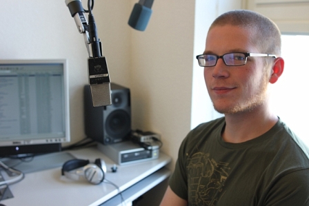 KSV-Spitzenkandidat David Lang mit kurzen Haaren, schwarzer Brille und grünem Shirt im FM4-Studie mit Mikrophon und Bildschirm im Hintergrund